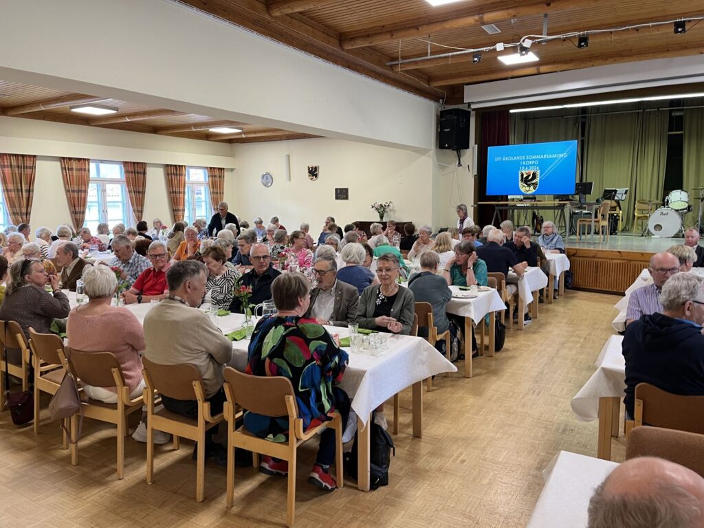 En stor grupp äldre vuxna sitter vid bord i en samlingslokal och pratar och äter. På en skärm längst fram i rummet visas text och ett vapen, och på scenen syns trummor och musikutrustning.