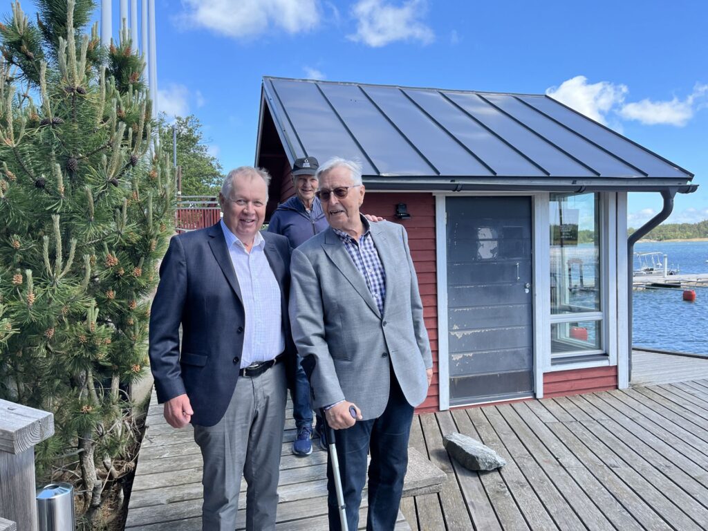 Tre äldre män står på en träbrygga nära en liten röd stuga med plåttak. Två av dem står i förgrunden och ler, medan en står bakom dem. I bakgrunden syns en sjö och båtar under en blå himmel.