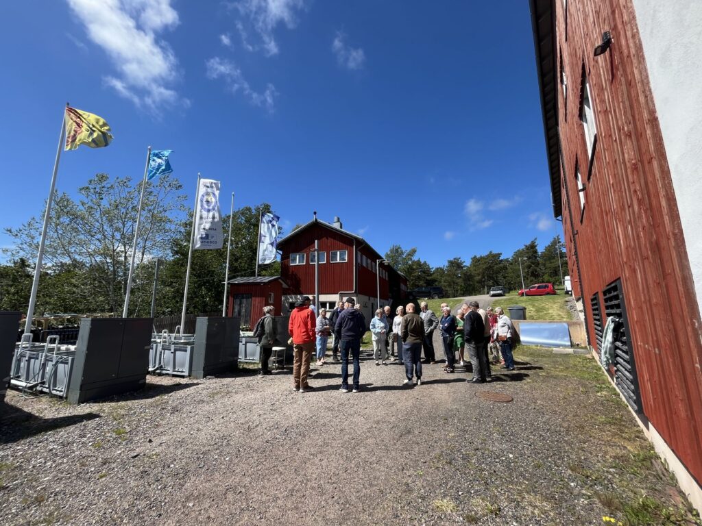 En grupp människor står utomhus på en grusväg nära röda träbyggnader och flera flaggstänger, under en klarblå himmel med spridda moln. Metallcontainrar syns längs sidorna.
