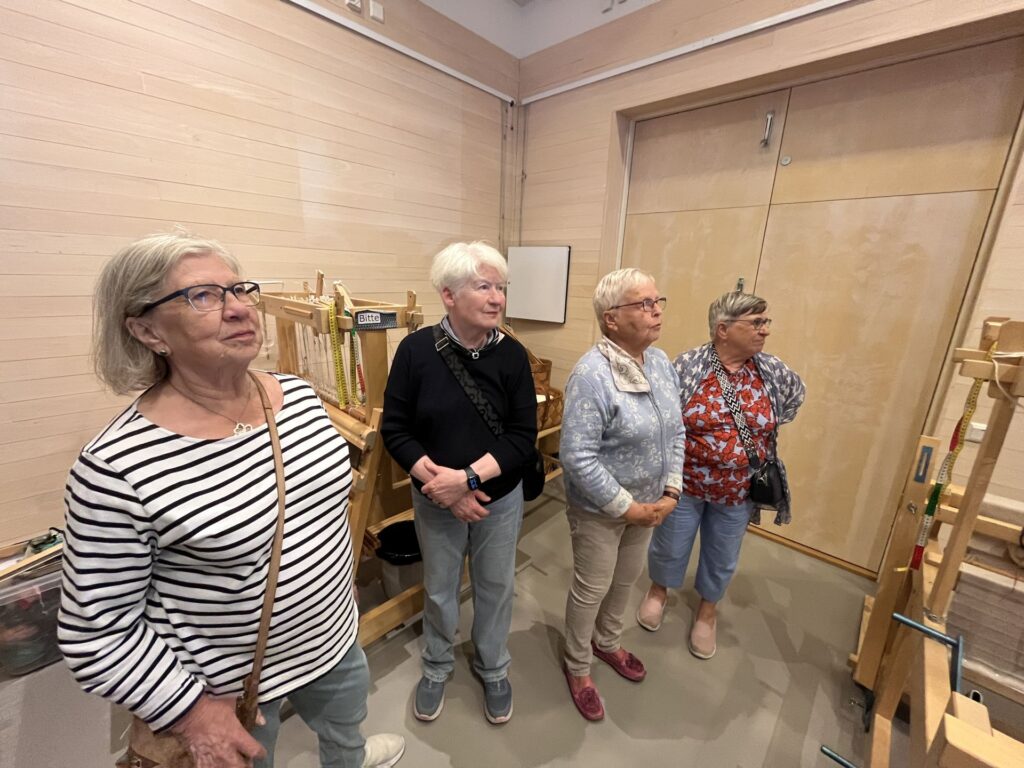 Fyra äldre kvinnor står inomhus vid vävstolar av trä och tittar åt samma håll. Rummet har ljusa väggar och en stängd trädörr. Kvinnorna är klädda i vardagskläder och verkar uppmärksamma.