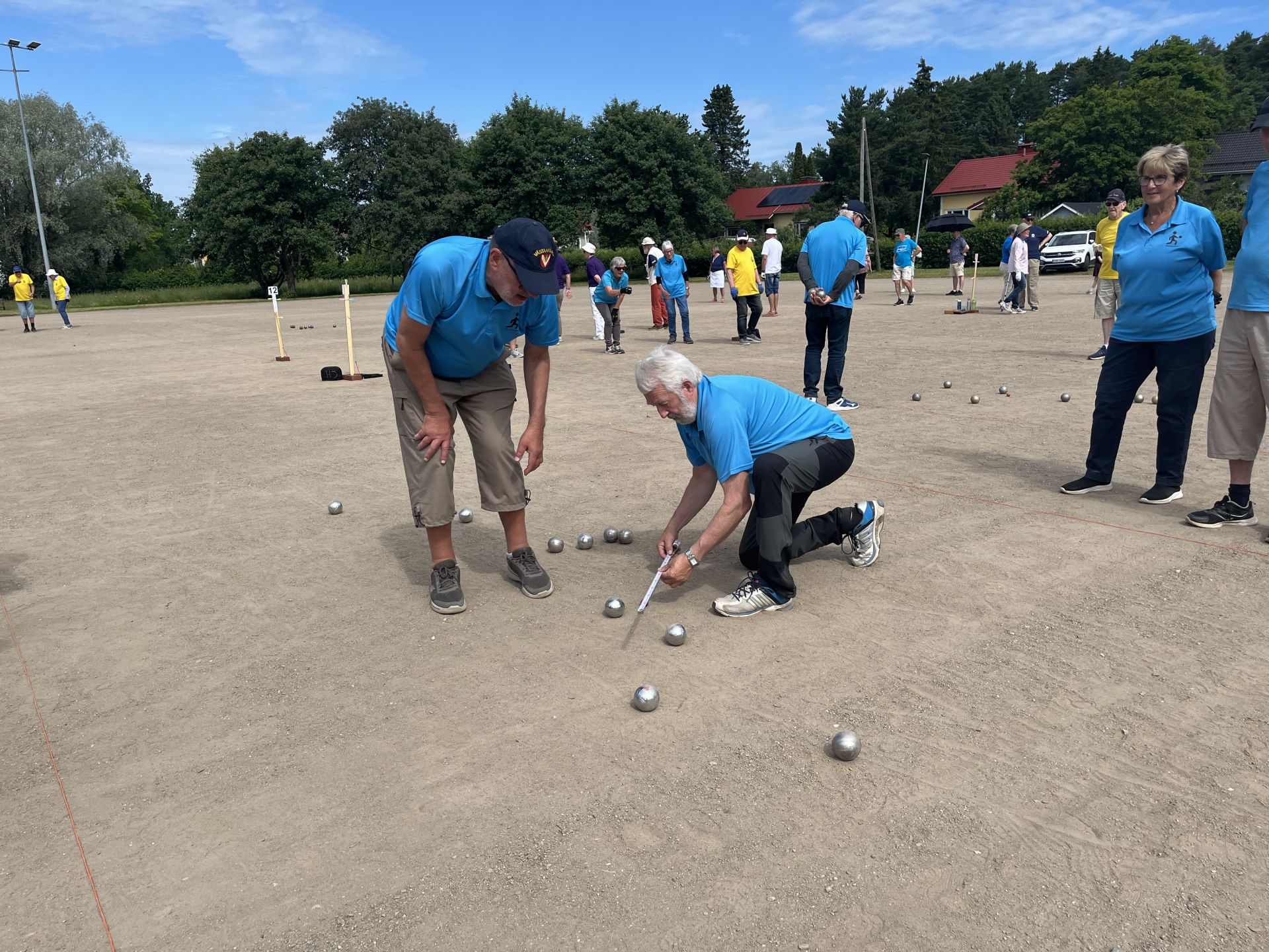 Flera personer spelar pétanque på en stor grusplan. Två spelare i blå tröjor är upptagna med att mäta avståndet mellan metallkulorna, medan andra står i bakgrunden. Bakom planen syns träd och hus.