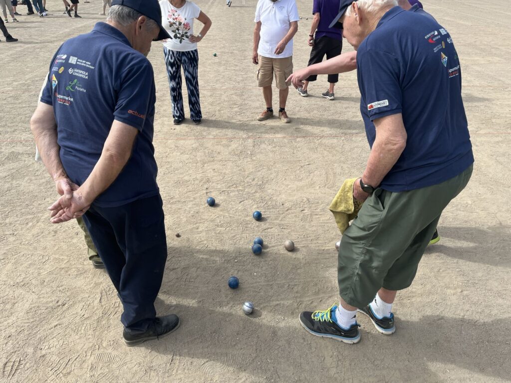 Flera personer står på en sandig yta och tittar på bocce- eller petanquekulor som ligger på marken; i förgrunden står två män i blå skjortor, varav den ene håller en handduk.