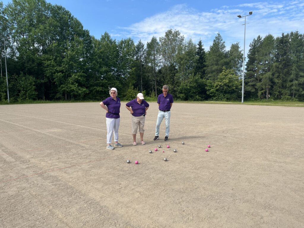 Tre personer i lila tröjor står på en sandig spelplan och tittar ner på flera metall- och rosa bollar som ligger utspridda på marken. I bakgrunden syns träd och en klarblå himmel.
