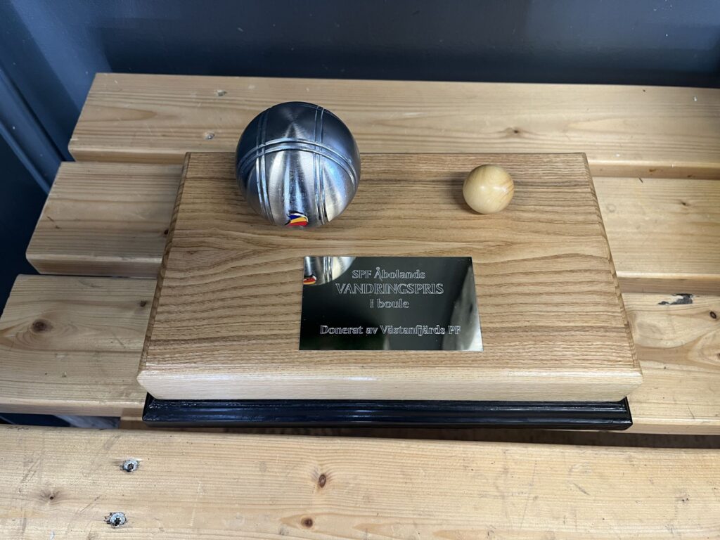 En pokal i trä med en silverfärgad metallkula och en liten gul kula ovanpå. På plaketten står det: SJF Åbneklub VANDREPRIS i boule Doneret af Vestamager/Isle FF. Pokalen står på en träbänk.