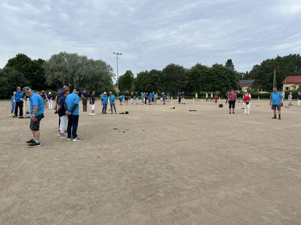 En stor grupp människor spelar pétanque på en grusplan, uppdelade i flera matcher. De flesta deltagarna bär blå, lila eller röda tröjor. I bakgrunden syns träd och byggnader under en molnig himmel.