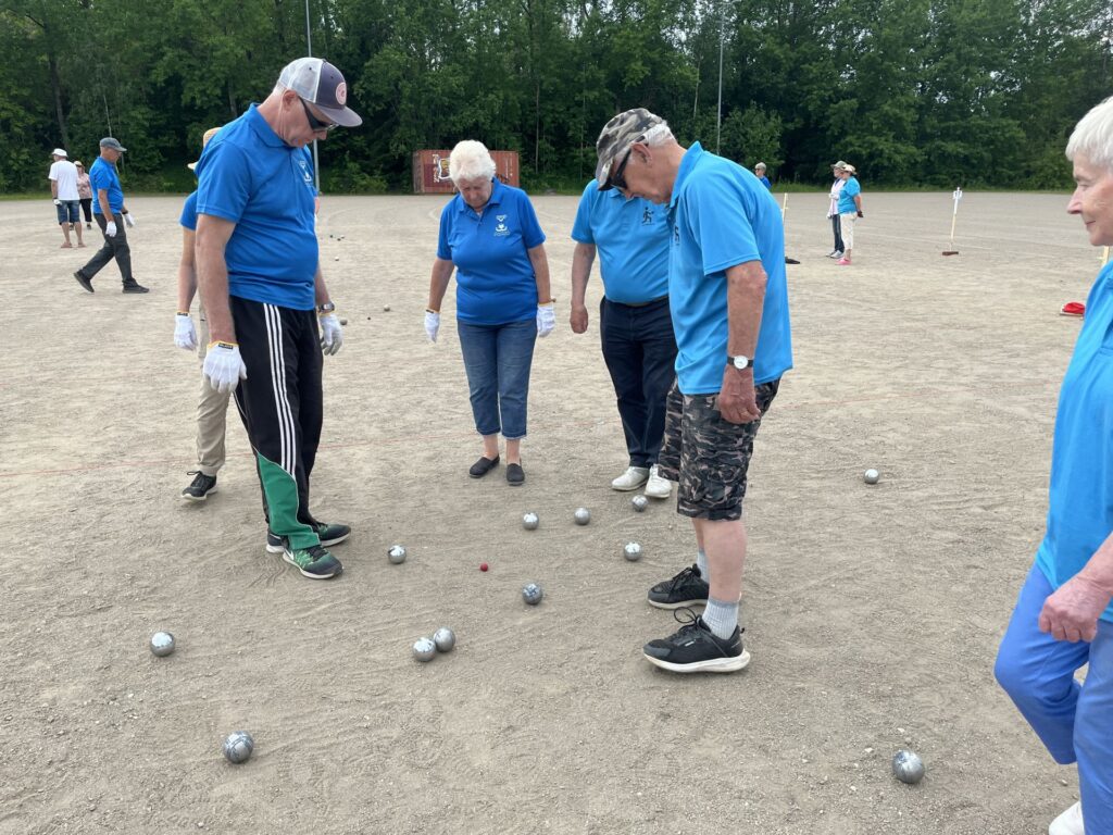 Flera äldre personer i blå tröjor står på en grusplan och tittar ner på metallkulor som ligger utspridda runt deras fötter; det ser ut som om de spelar pétanque eller boccia. I bakgrunden syns träd och fler spelare.
