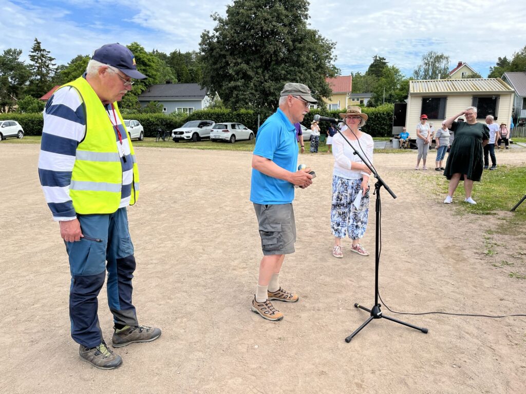 Tre äldre personer står utomhus nära en mikrofon. Mannen till vänster har på sig en gul väst och en keps, mannen i mitten är klädd i blått och kvinnan till höger har en hatt på sig. I bakgrunden syns människor och bilar.