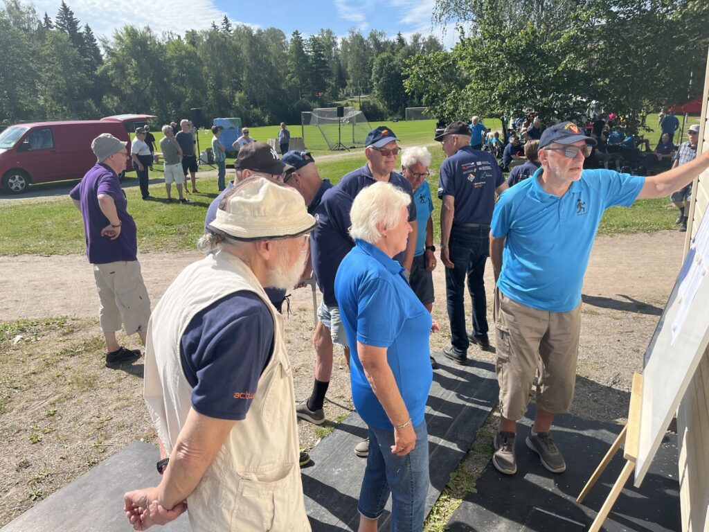 En grupp äldre personer, varav några bär blå skjortor och kepsar, står utomhus och tittar på en informationsskylt. Andra sitter eller står i närheten på en solig dag på en gräsplan med träd i bakgrunden.