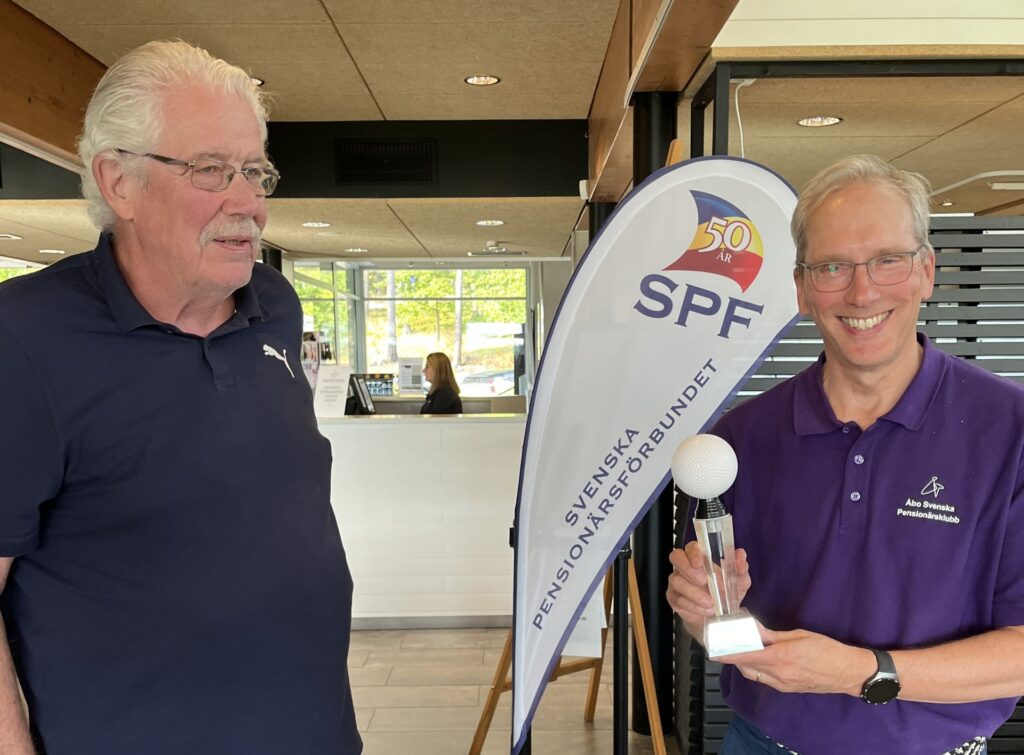 Två äldre män står inomhus bredvid en banderoll med SPF:s logotyp. Den ene mannen håller i en pokal i form av en golfboll på ett stativ och ler. Den andre mannen tittar på honom. I bakgrunden syns en receptionsdisk.