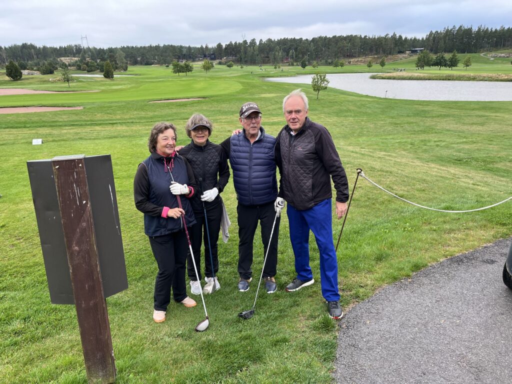 Fyra vuxna klädda i golfkläder står på en golfbana med klubbor i händerna. Bakom dem syns grönt gräs, träd, en damm och en molnig himmel. I förgrunden syns en vägskylt och en asfalterad stig.