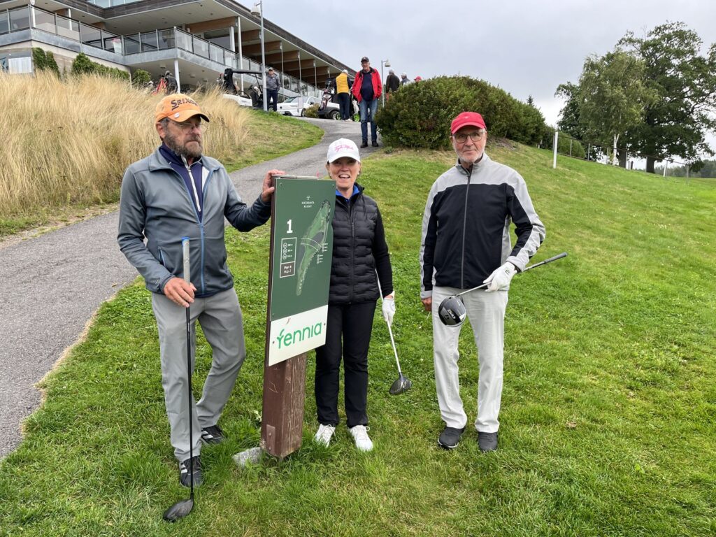 Tre vuxna i golfkläder står på gräset nära en golfbaneskylten med siffran ”1” och texten ”Fennia”. Två av dem håller i golfklubbor. I bakgrunden syns en byggnad, en stig och flera personer.