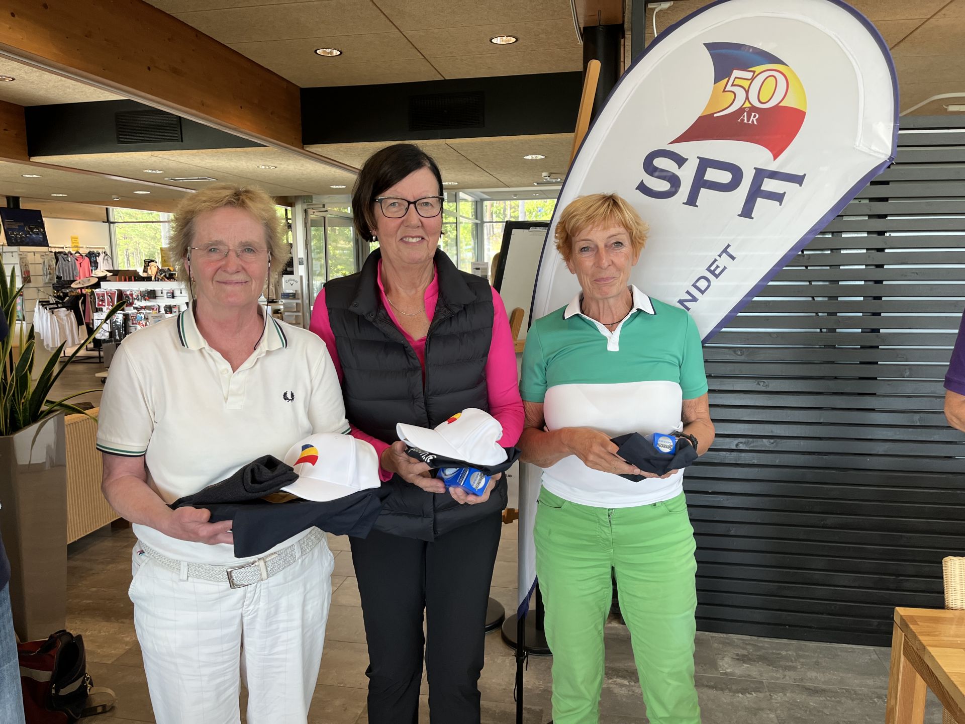 Tre kvinnor står inomhus och håller i golfkepsar och priser. De står framför en vit banderoll med texten ”SPF 50 ÅR”. I bakgrunden syns hyllor och en disk med varor.