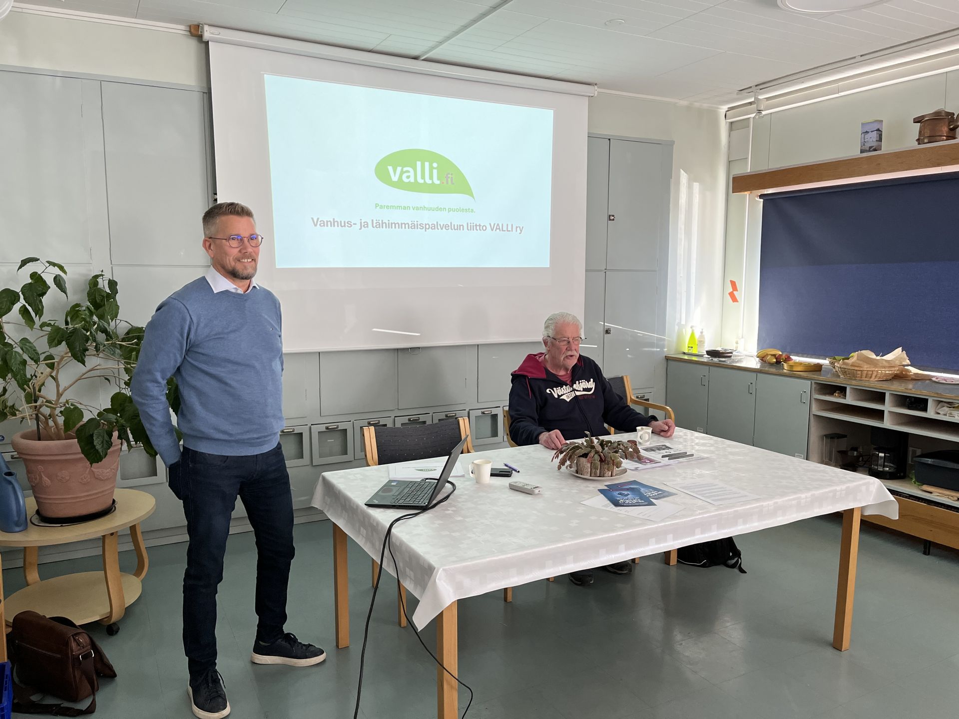 Två män befinner sig i ett rum med ett bord och en projicerad presentation på väggen. Den ena mannen står och talar, medan den andra sitter vid bordet med papper. Presentationsbilden visar logotypen för VALLI.fi och information på finska.