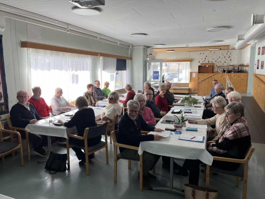 En grupp äldre vuxna sitter runt bord i ett ljust rum, engagerade i diskussioner eller pappersarbete. Några är vända mot kameran, andra har ryggen vänd. Papper och pennor ligger synliga på borden.