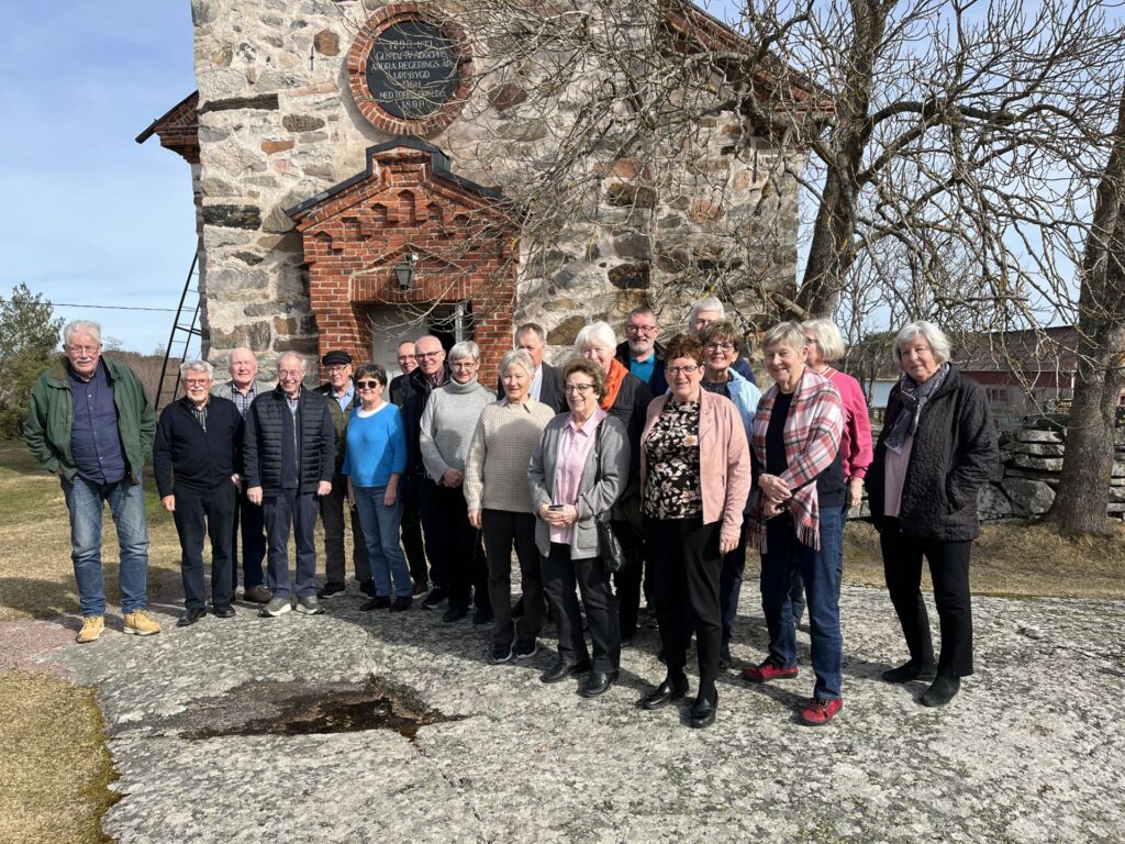 En grupp på cirka 20 vuxna står utanför en stenbyggnad med en ingång av tegel. De poserar för ett foto en solig dag, med lövlösa träd och en klar himmel i bakgrunden.