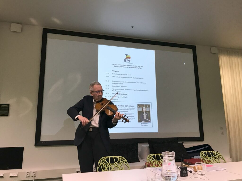En man i kostym spelar fiol framför en projektionsskärm i ett konferensrum. I förgrunden står ett bord med drycker och papper. På presentationsbilden syns SPF:s logotyp och evenemangets dagordning.