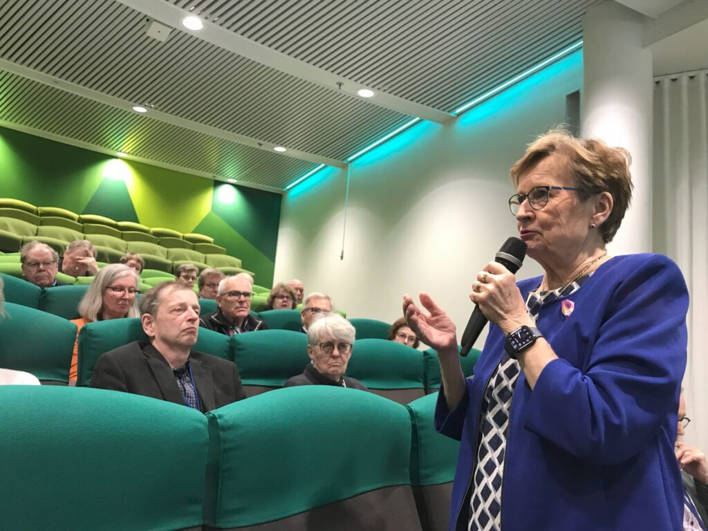 En kvinna i en blå jacka talar i en mikrofon framför en publik som sitter i gröna stolar i ett starkt upplyst rum med ett grönt och vitt tak. Publiken består främst av äldre vuxna som lyssnar uppmärksamt.