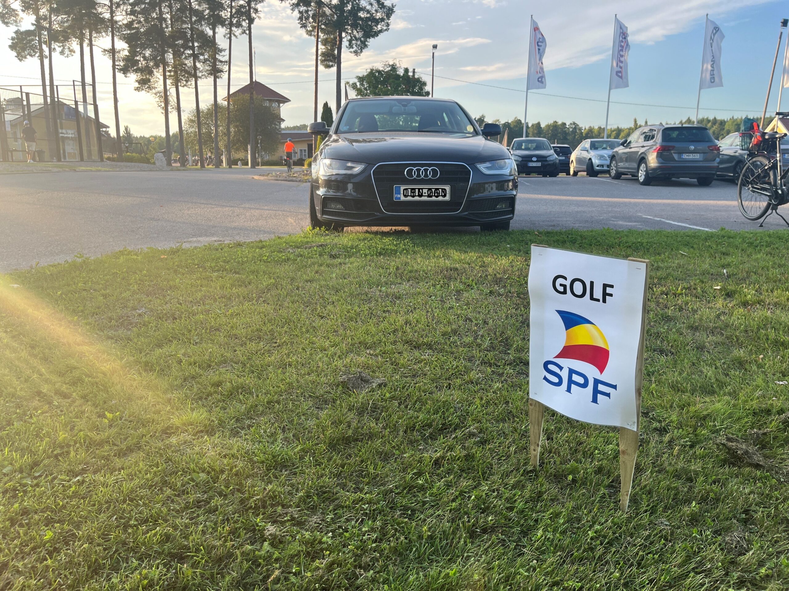 En svart Audi står parkerad på gräset intill en väg. Framför bilen står en liten skylt med texten ”GOLF SPF” och en färgglad logotyp. I bakgrunden syns flera bilar och cyklar.