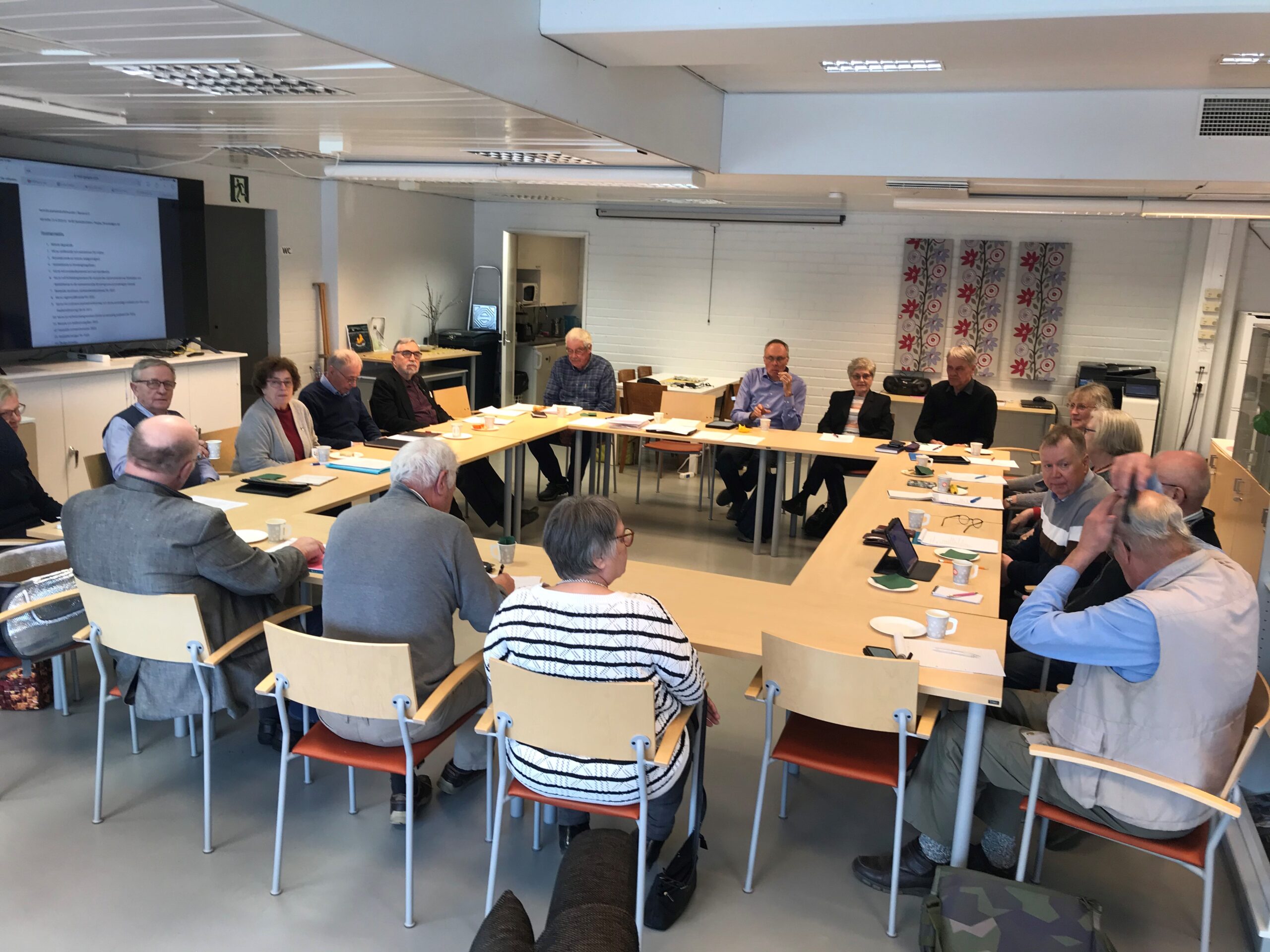 En grupp människor sitter runt ett stort, U-format bord i ett mötesrum och diskuterar. Vissa har papper och kaffekoppar framför sig, och en presentation visas på en skärm i bakgrunden.