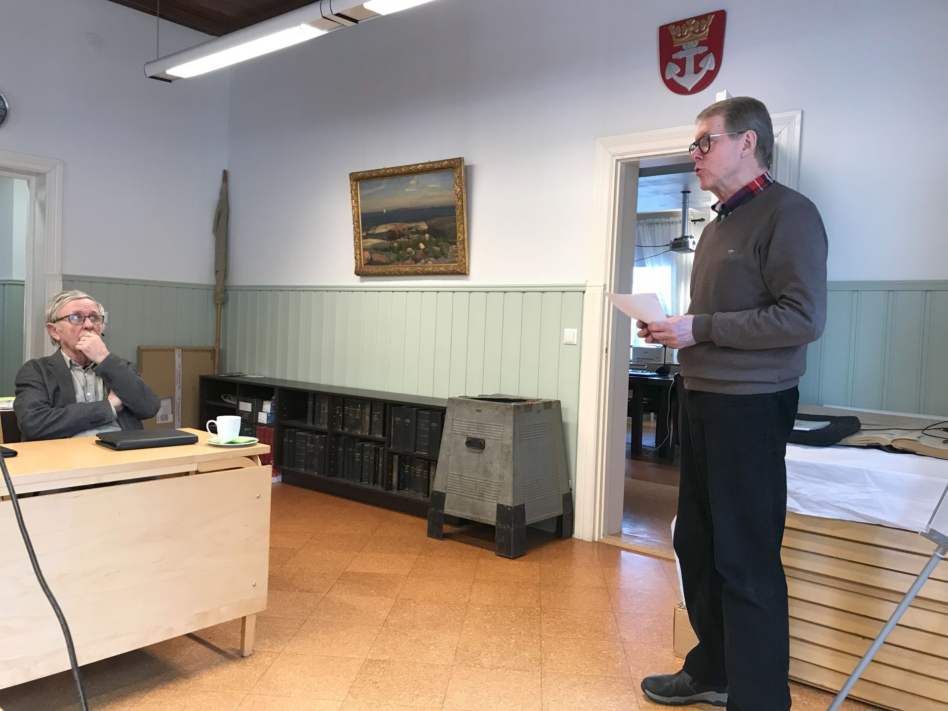 En man står och läser ur en tidning medan en annan man sitter vid ett skrivbord med en bärbar dator och en kopp i ett rum med gröna paneler, bokhyllor och en tavla på väggen.