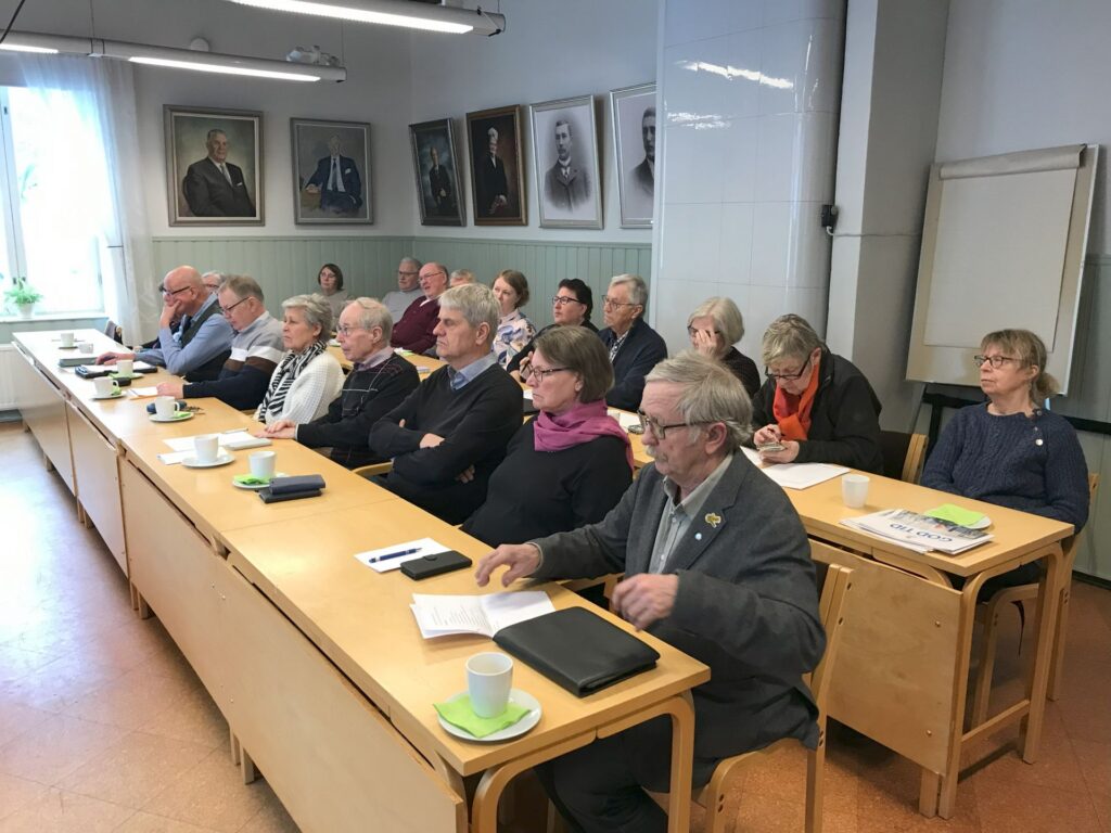 En grupp äldre vuxna sitter vid skrivbord i ett klassrum och lyssnar uppmärksamt. På skrivborden ligger anteckningsböcker, papper och muggar. På väggen bakom dem hänger porträtt av män, och i hörnet står ett blädderblock.