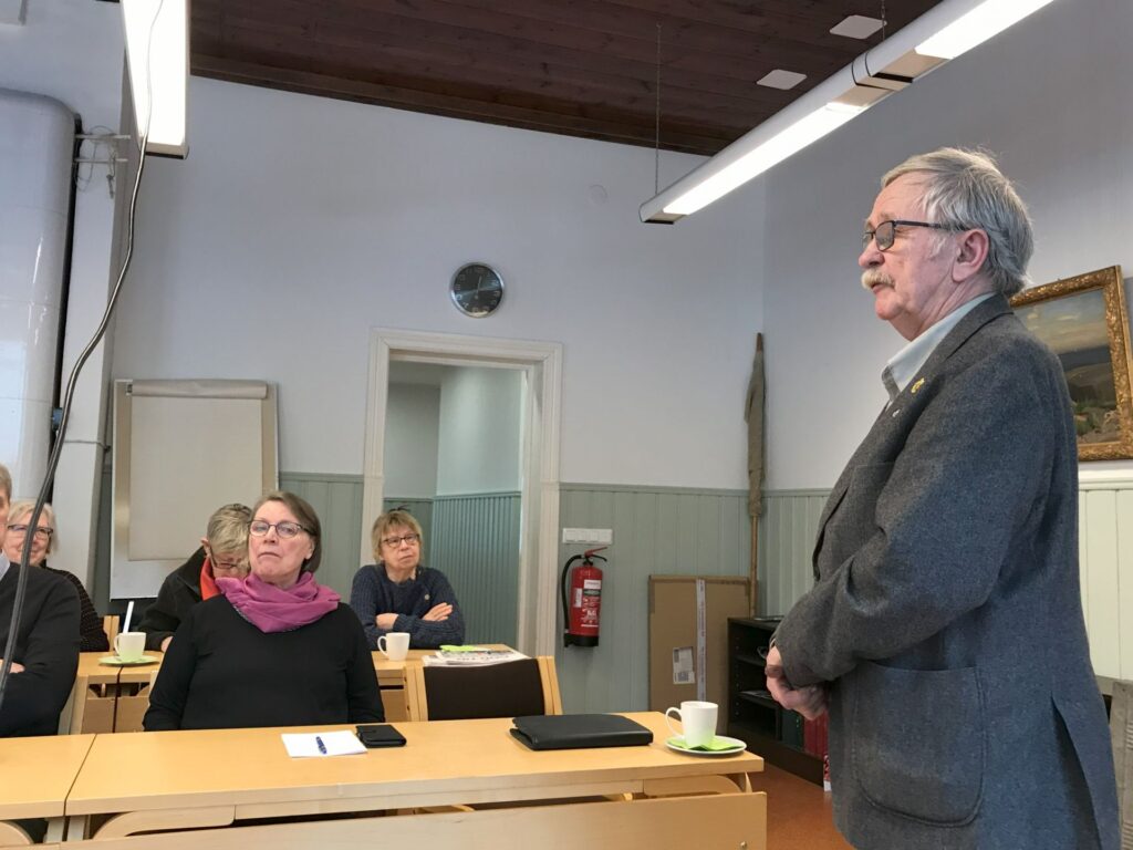 En man i grå kostym står och talar till en liten grupp som sitter vid skrivbord i ett klassrum. Flera personer lyssnar uppmärksamt. I rummet syns muggar, anteckningsböcker och en brandsläckare.