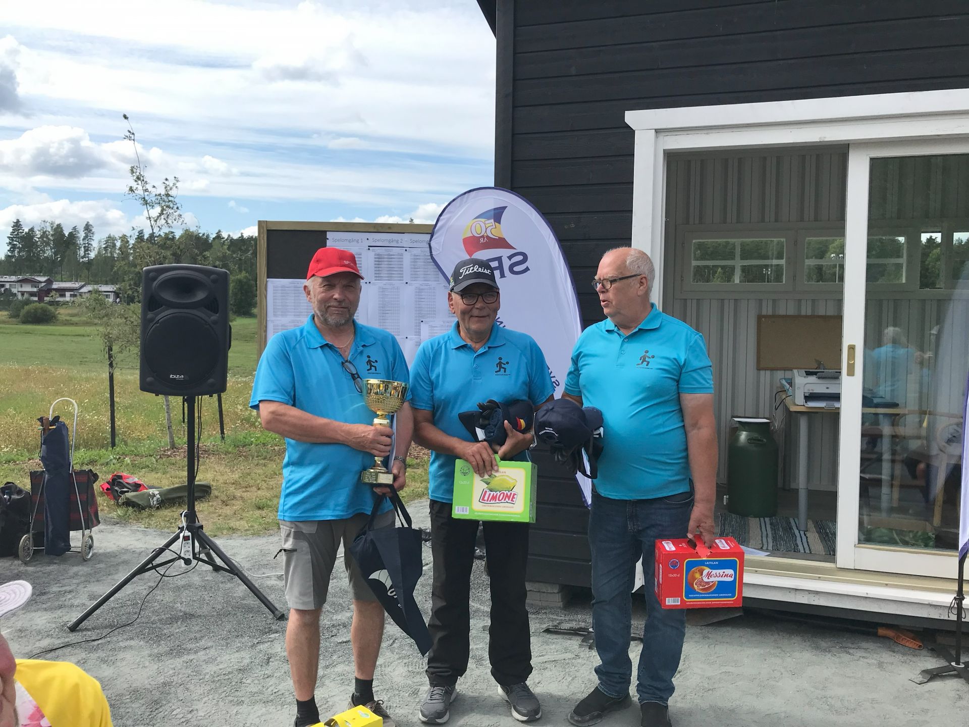 Tre män klädda i likadana blå skjortor står framför en byggnad. En av dem håller i en pokal och ett paraply, en annan håller i en låda och ett pris, och den tredje håller i en låda. I bakgrunden syns en högtalare och en banderoll.