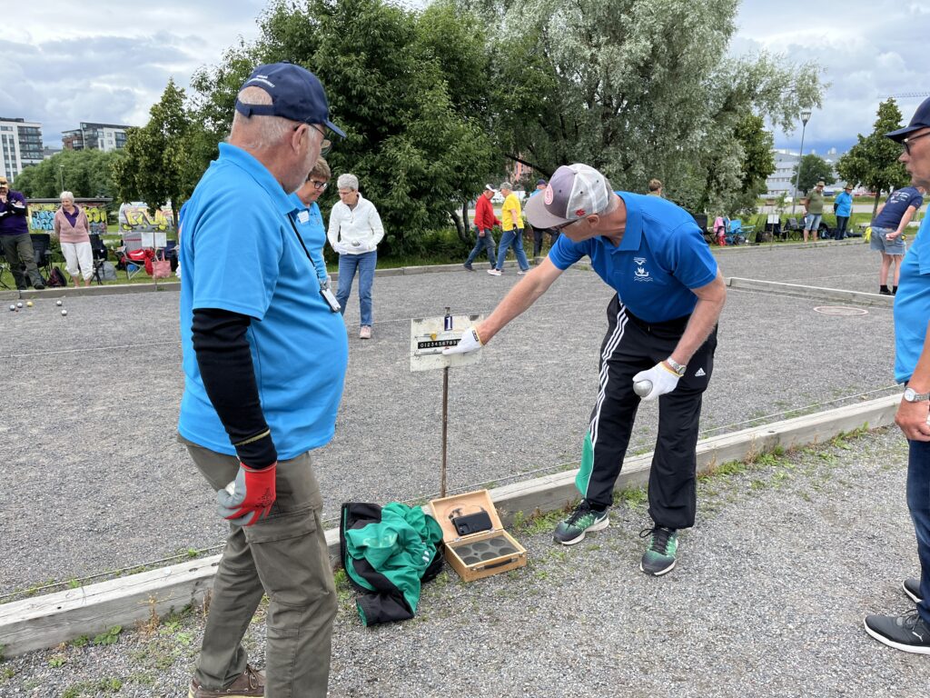 Två äldre män i blå skjortor undersöker utrustningen på en petanquebana med grusunderlag. En av dem pekar på ett metallstativ med en mätanordning. I bakgrunden spelar eller tittar andra på under en molnig himmel. Träd och byggnader syns i bakgrunden.