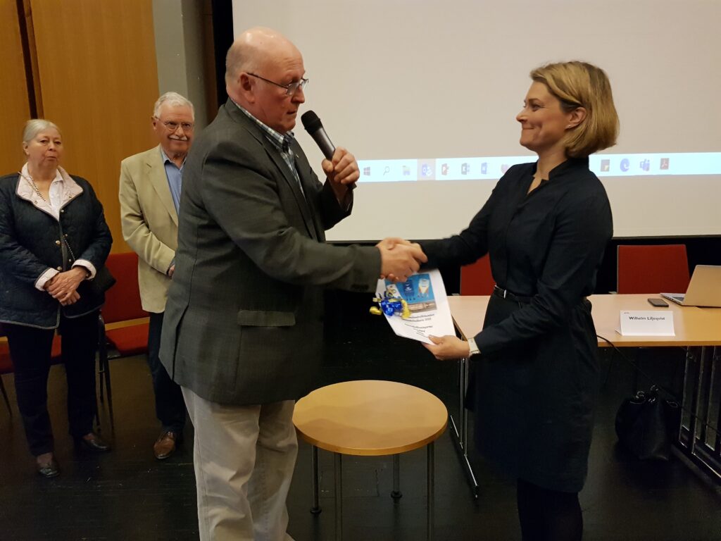 En man i kavaj skakar hand med en kvinna i en mörk klänning samtidigt som han håller i en mikrofon och ett certifikat. Två personer står i bakgrunden, och till höger syns ett bord med stolar.