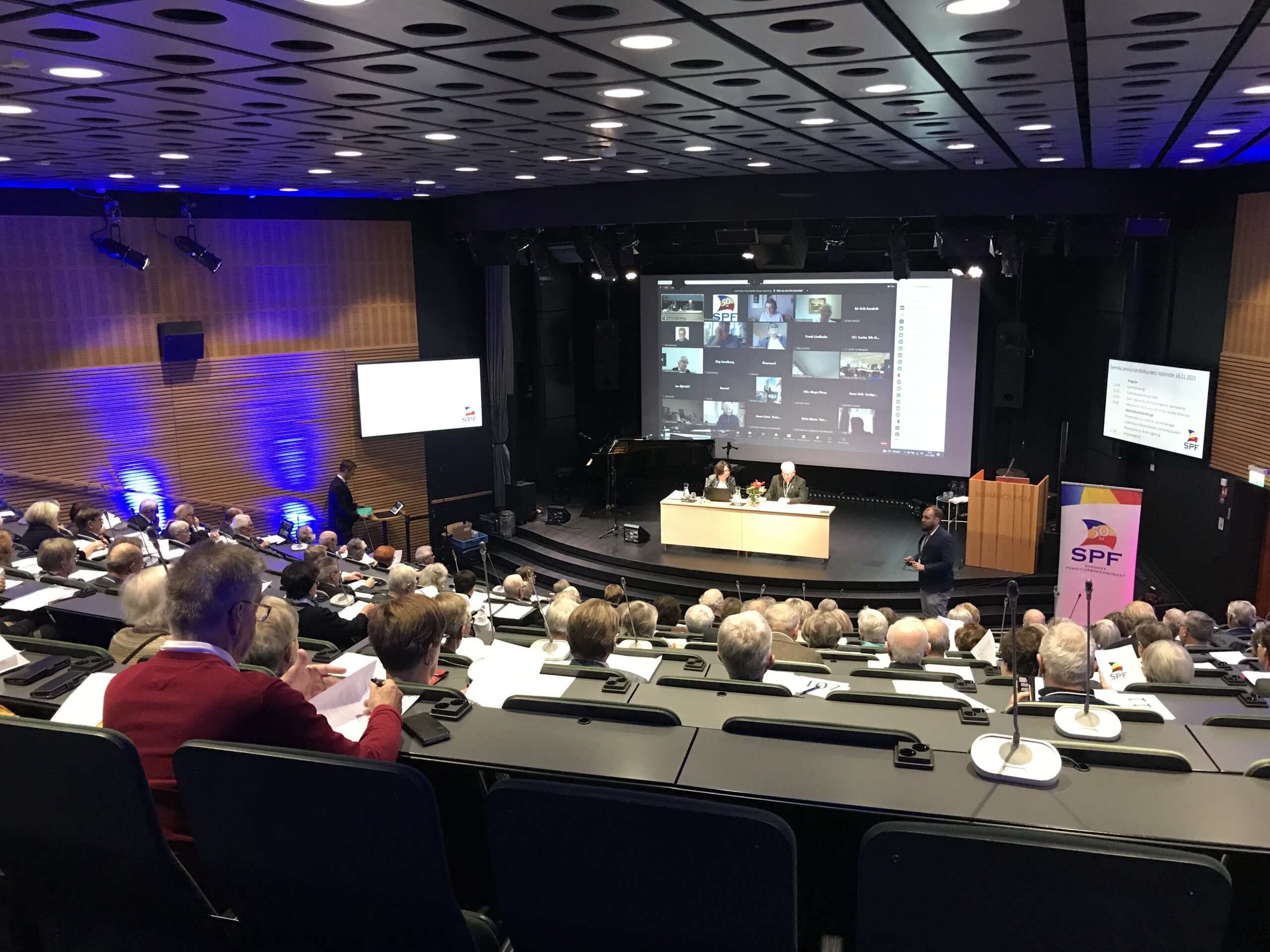 Ett konferensrum med rader av sittande deltagare som vetter mot en scen, där två personer sitter vid ett bord framför en stor skärm som visar ett videosamtal med flera deltagare. SPF:s logotyp syns på ett podium i närheten.