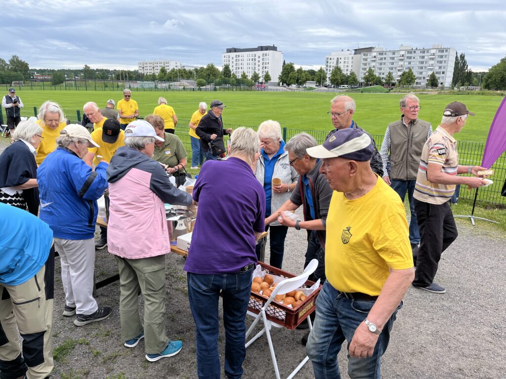 En grupp äldre människor, varav några bär gula tröjor, samlas utomhus runt bord med mat och brickor med brödbullar. I bakgrunden syns bostadshus och en gräsbevuxen äng under en molnig himmel.