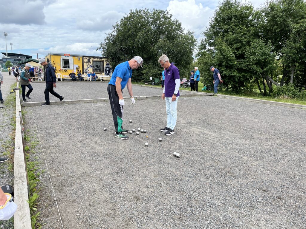 Två män står på en grusbelagd pétanquebana och tittar på silverfärgade bollar som ligger på marken. I bakgrunden syns flera personer, en gul husvagn och träd under en molnig himmel.
