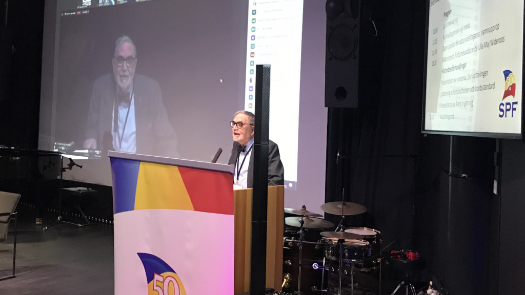 En person står vid ett talarstativ som är dekorerat med en 50-årsjubileumslogotyp och SPF-logotypen och talar till publiken. På en projektionsskärm bakom visas ett videosamtal och presentationsbilder. I bakgrunden syns musikinstrument.