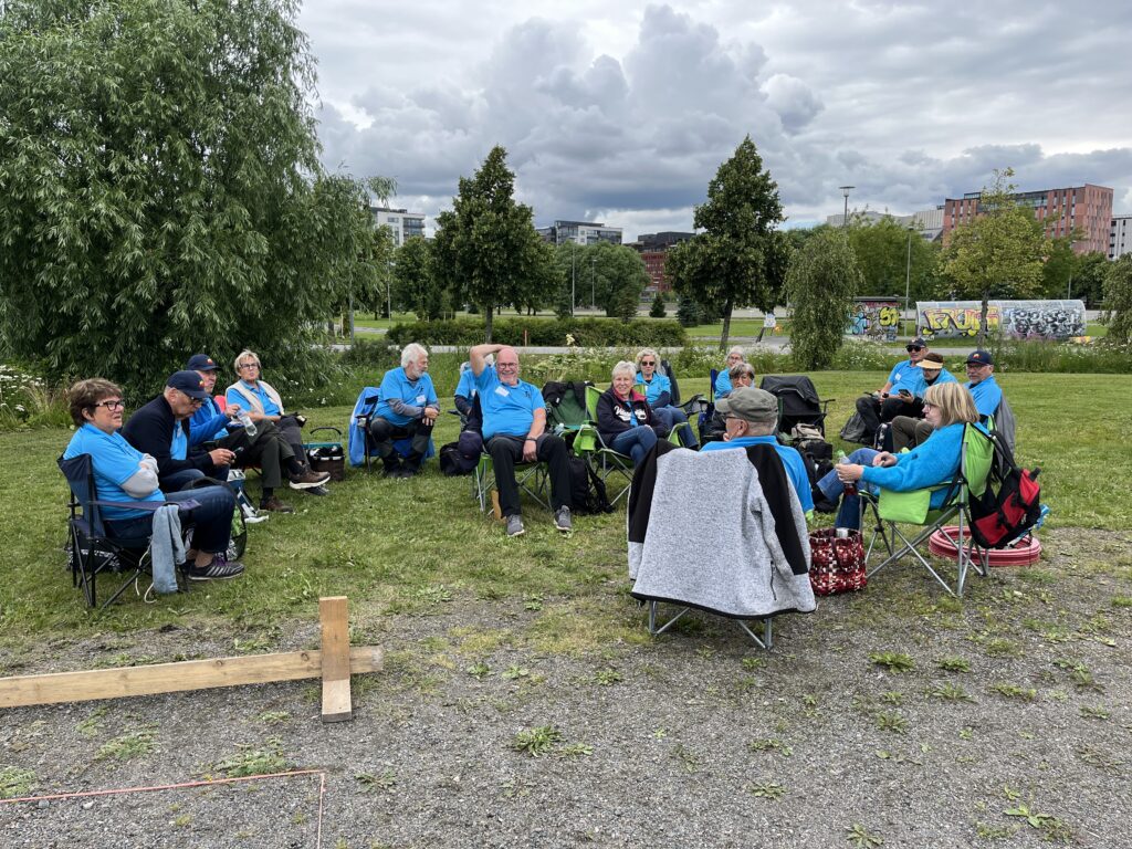 En grupp människor i likadana blå tröjor sitter på campingstolar i en gräsbevuxen park och pratar och kopplar av. I bakgrunden syns träd, byggnader och en molnig himmel, och det ligger väskor och jackor utspridda runt omkring.