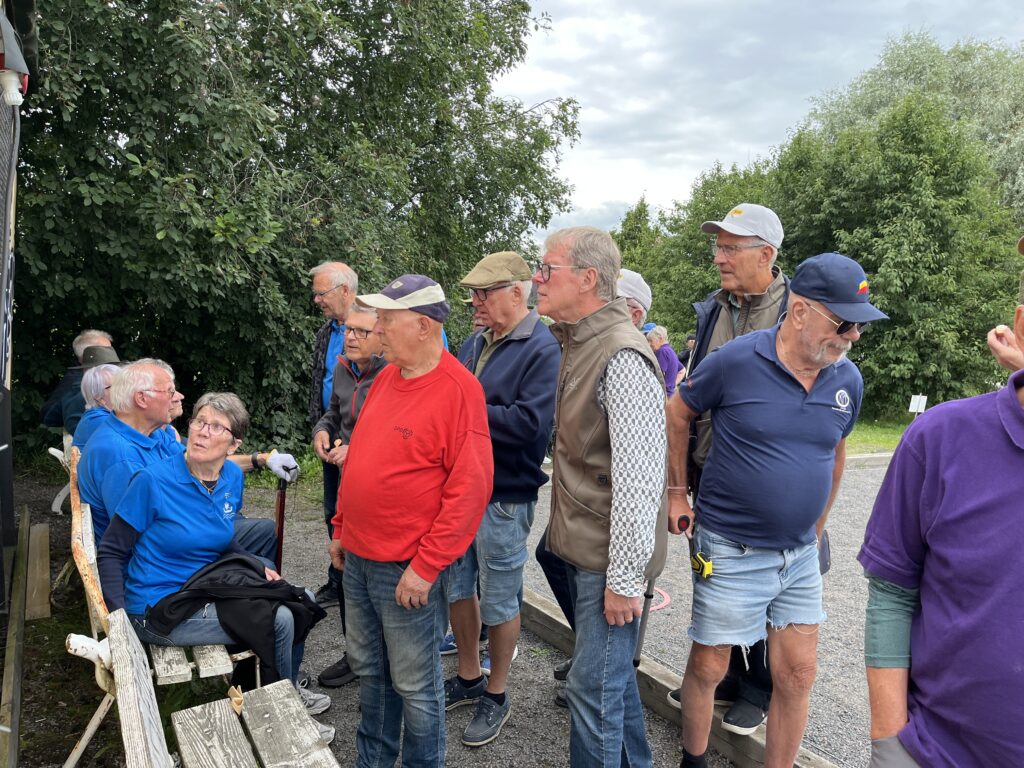 En grupp äldre personer, främst män, står och sitter utomhus nära träd och en grusgång. Vissa bär hattar och glasögon, och de flesta är klädda i vardagskläder, bland annat shorts och jackor.