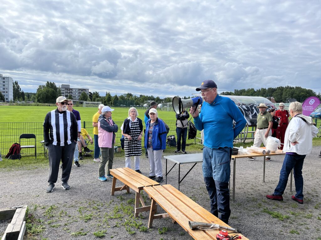 En grupp människor står utomhus nära bänkar och bord på en grusgång. En man talar i en megafon. I bakgrunden syns en gräsbevuxen äng, träd och flera byggnader under en molnig himmel.