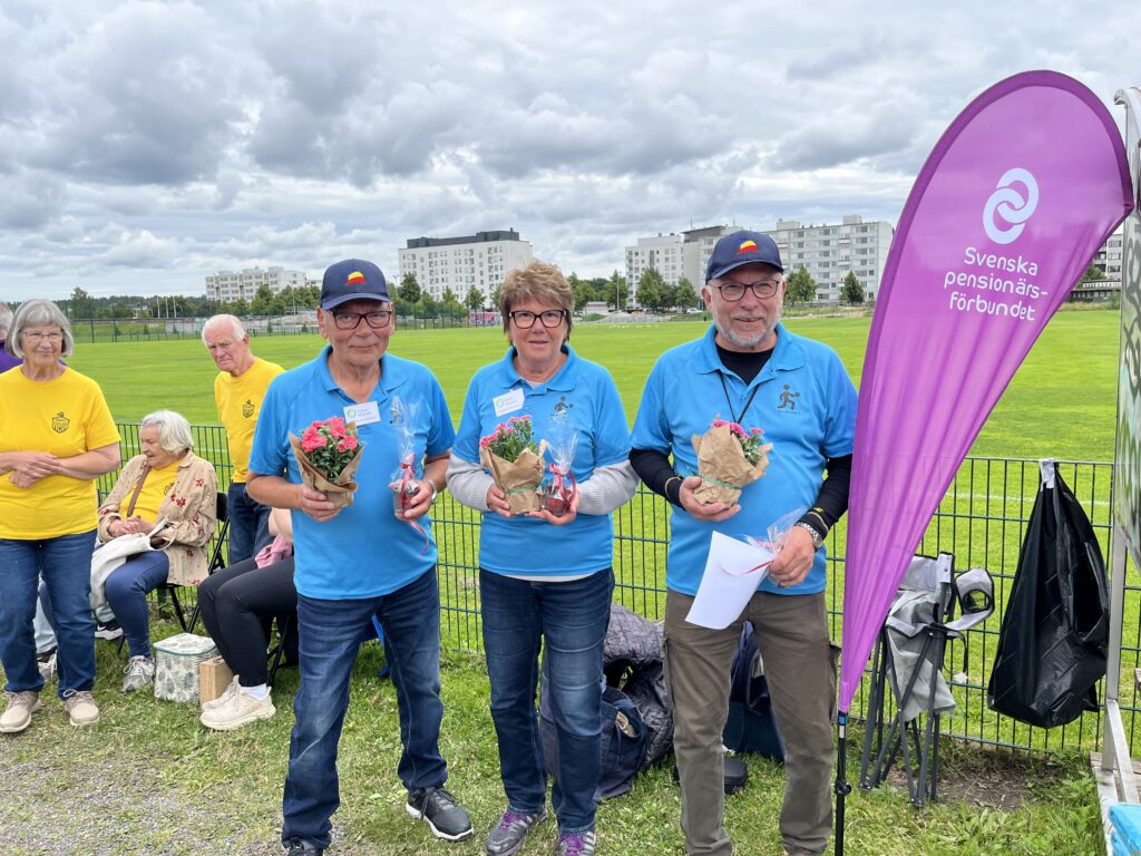 Tre äldre personer i blå skjortor står med blomsterbuketter i händerna bredvid en lila flagga från Svenska pensionärsförbundet utomhus, medan andra människor sitter i bakgrunden där det syns gräsbevuxna fält och byggnader.