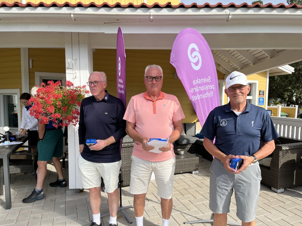 Tre äldre män står sida vid sida med små lådor i händerna framför en gul byggnad med lila banderoller. De är klädda i avslappnad golfklädsel och bär solglasögon. I närheten hänger en blomkorg och det står utemöbler.