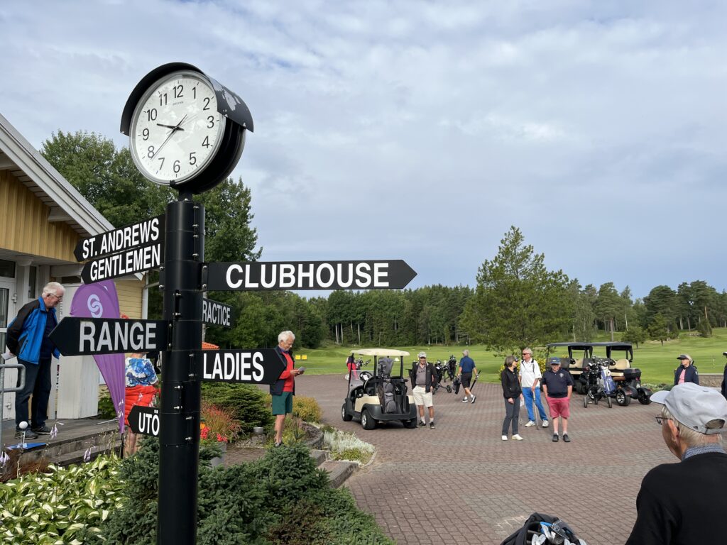 En scen på en golfbana där människor går bredvid golfbilar, med en byggnad till vänster och en svart vägvisare med skyltar som pekar mot ”Clubhouse”, ”Range”, ”St. Andrews”, ”Gentlemen” och ”Ladies” under en klocka som visar 10:15.