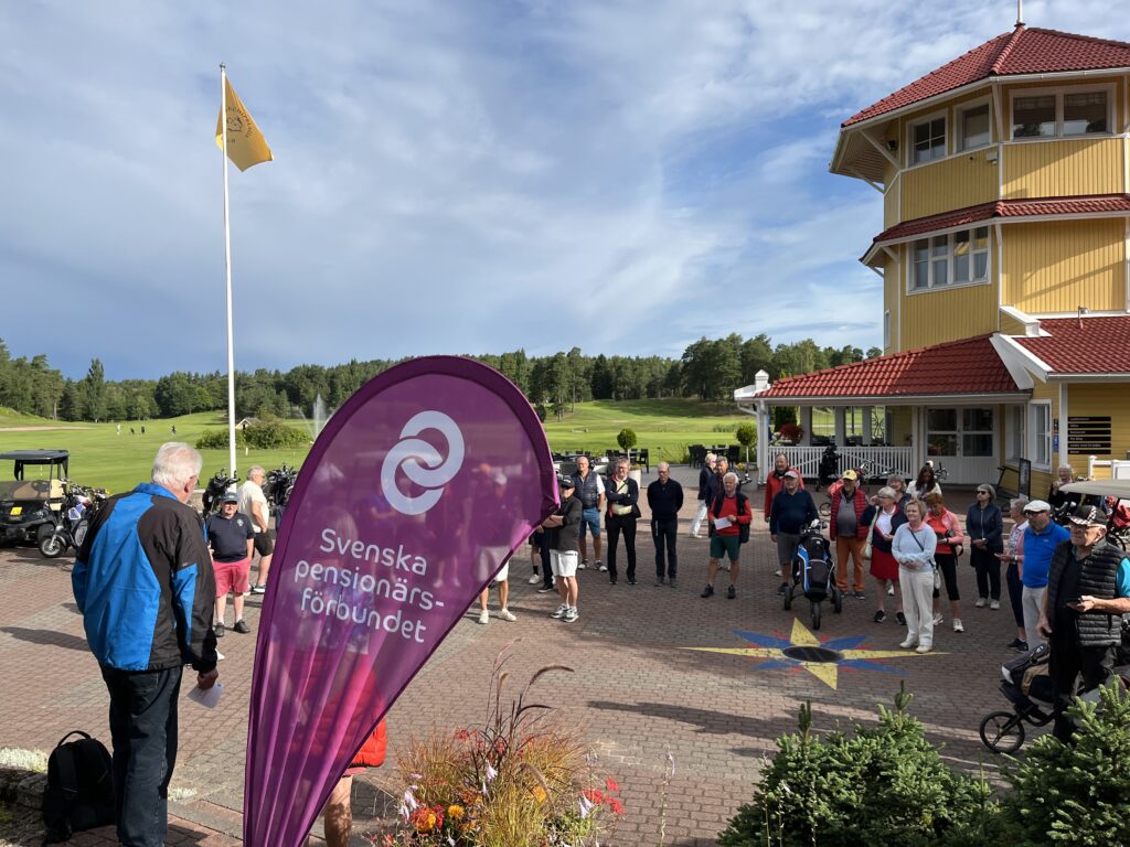 En grupp människor står utanför en gul byggnad med röda tak, samlade nära en lila flagga från Svenska pensionärsförbundet. En man talar till folkmassan. I bakgrunden syns en golfbana och en flagga under en blå himmel.