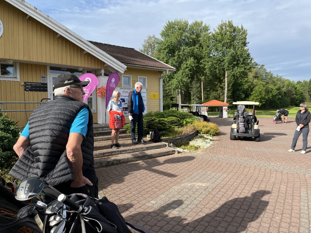 En grupp människor står utanför en gul träbyggnad bredvid en golfbil. En person lutar sig mot en golfväska i förgrunden. I bakgrunden syns träd och en blå himmel.