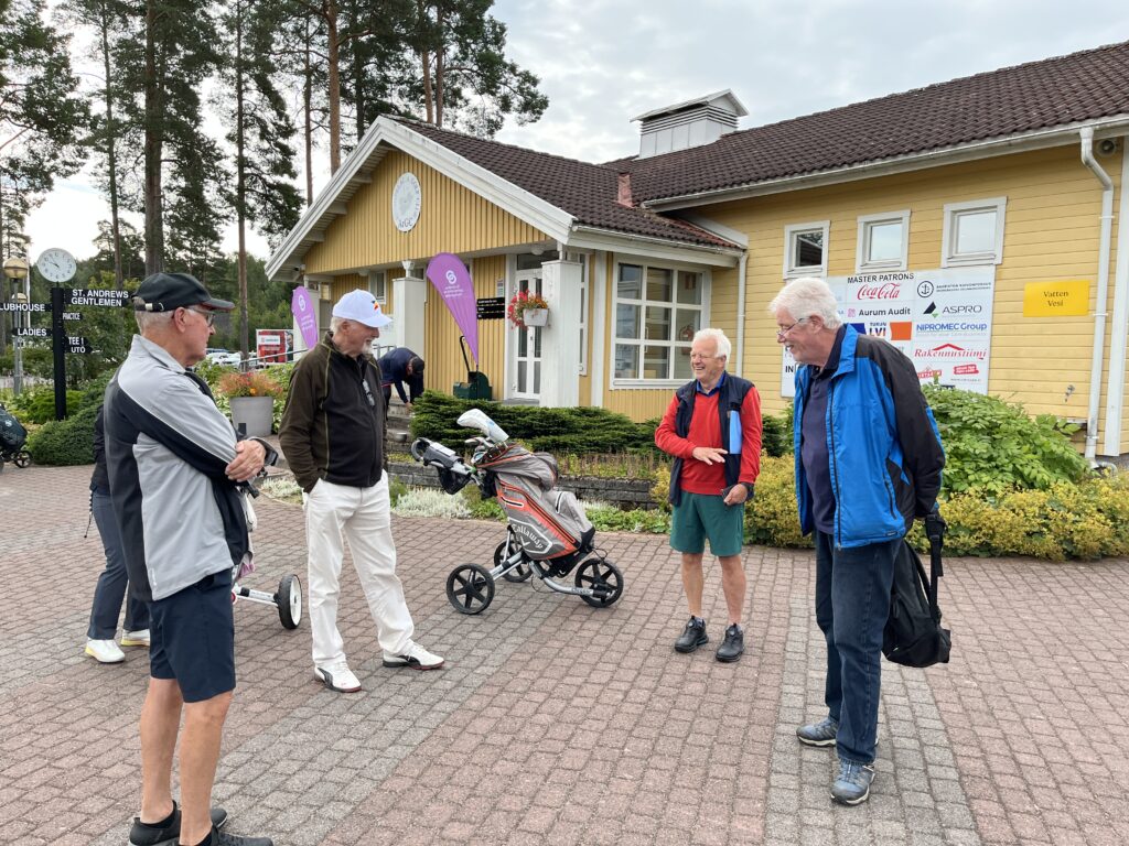 Fem äldre män står och pratar nära en gul byggnad, där man kan se sponsorskyltar, golfväskor och grönska. En av männen har shorts på sig, medan de andra bär jackor eller golfkläder. Scenen verkar utspela sig vid ett golfevenemang.
