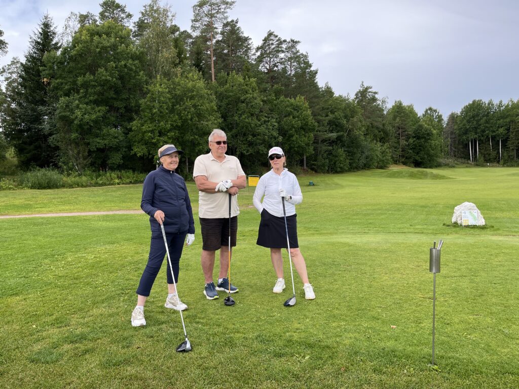 Tre personer i golfkläder står på en grön golfbana med golfklubbor i händerna. I bakgrunden syns träd och gräs. Det är molnigt, och golfutrustning syns till höger i bilden.