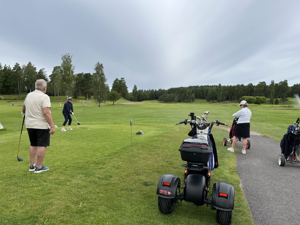 Tre personer befinner sig på en golfbana; en förbereder sig för att slå ut, en står i närheten och en annan står bredvid en golfbil och en elrullstol på vägen. Träd och grönt gräs fyller bakgrunden.