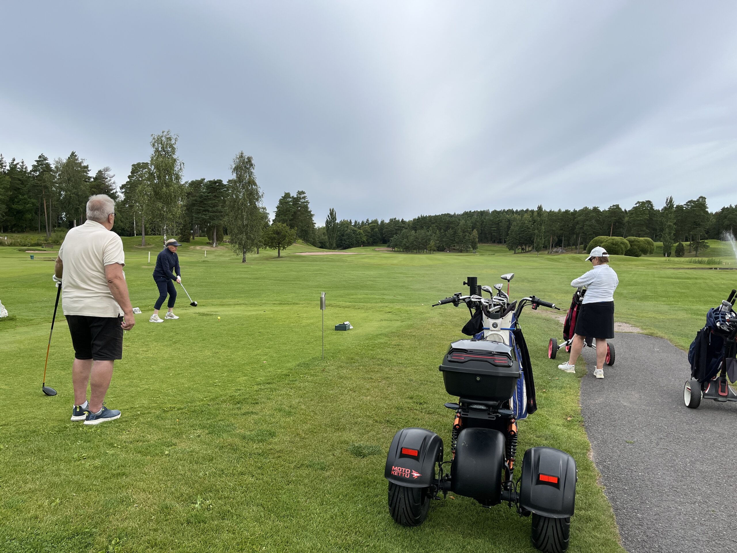Tre personer befinner sig på en golfbana; en förbereder sig för att slå ut, en står i närheten och en annan står bredvid en golfbil och en elrullstol på vägen. Träd och grönt gräs fyller bakgrunden.