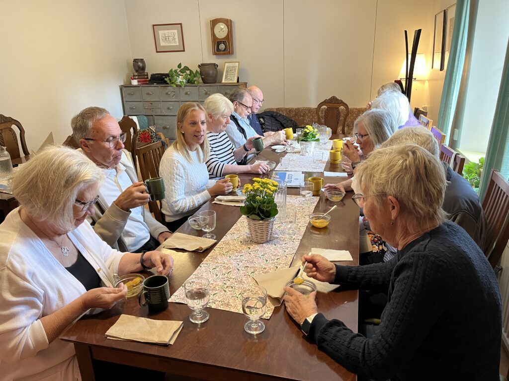 En grupp äldre sitter runt ett matbord i trä och äter och dricker. Bordet är dukat med skålar, muggar, blommor och servetter. Rummet är inrett med växter, tavlor och vintage-möbler.