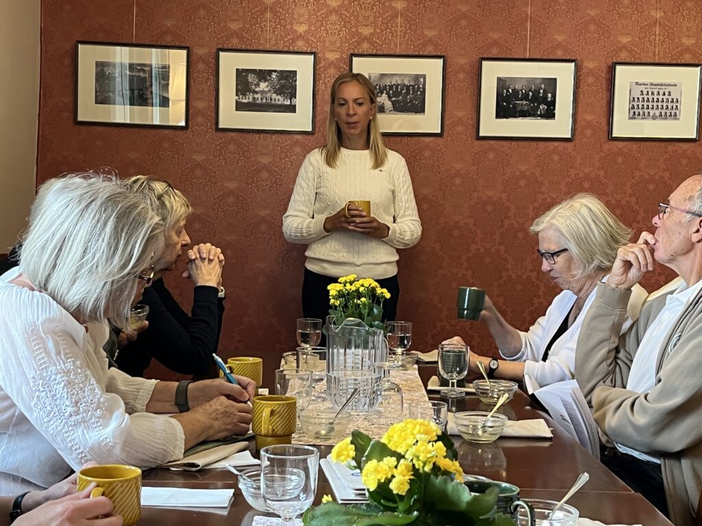 En grupp äldre vuxna sitter runt ett bord med gula blommor, dricker och skriver, medan en kvinna i en vit tröja står där med en mugg i handen. Svartvita foton hänger på den röda tapetserade väggen bakom henne.