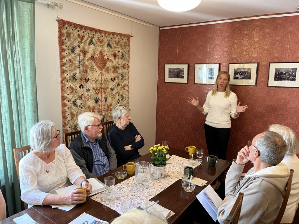 En kvinna står och talar till sex äldre personer som sitter i ett litet, mysigt inrett rum. I mitten står ett bord med koppar, en kanna och blommor. På väggarna hänger foton och en vävd matta.
