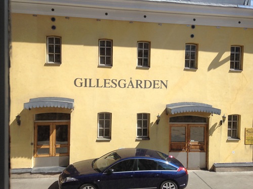 En gul byggnad med texten ”GILLESGÅRDEN” på fasaden, fyra fönster ovanför och fyra dörrar nedanför, en mörk bil parkerad framför på gatan samt två markiser över ingångsdörrarna.